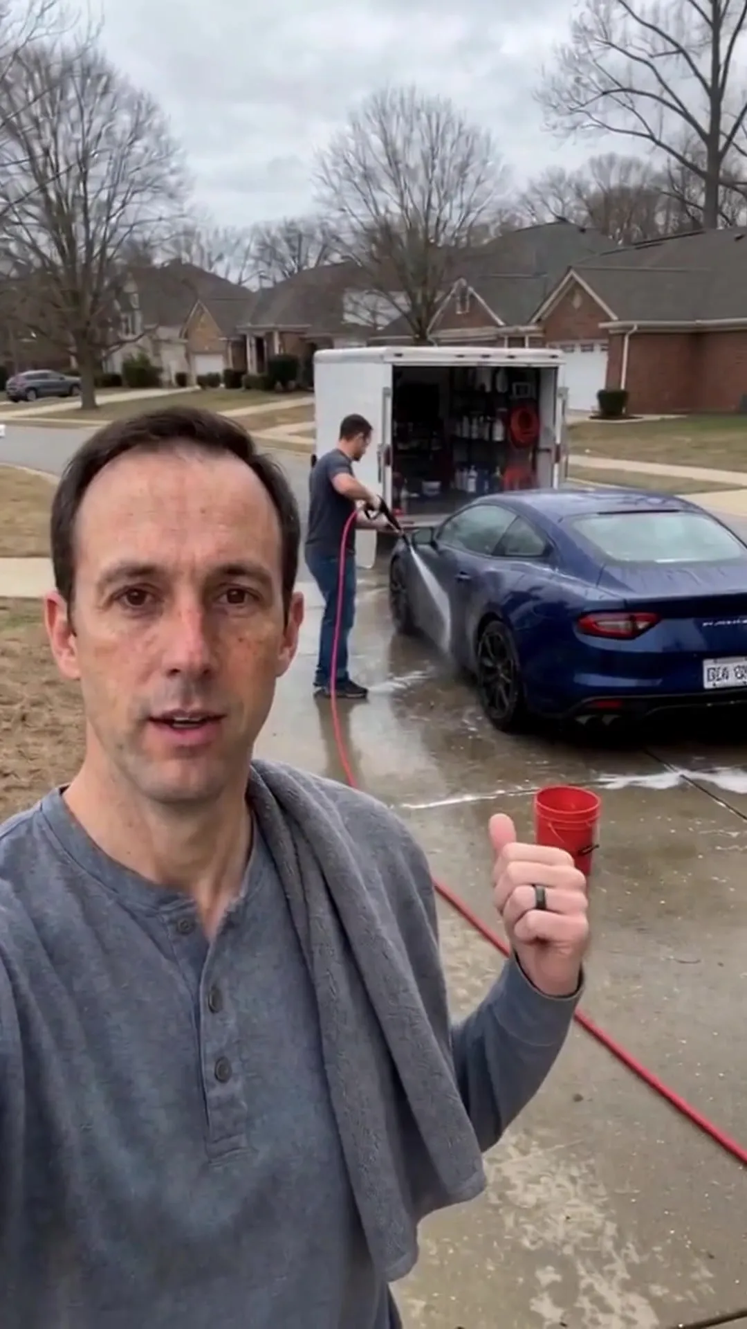 A crew member rinsing a blue car on a wet driveway beside an open trailer, a red bucket on the ground.