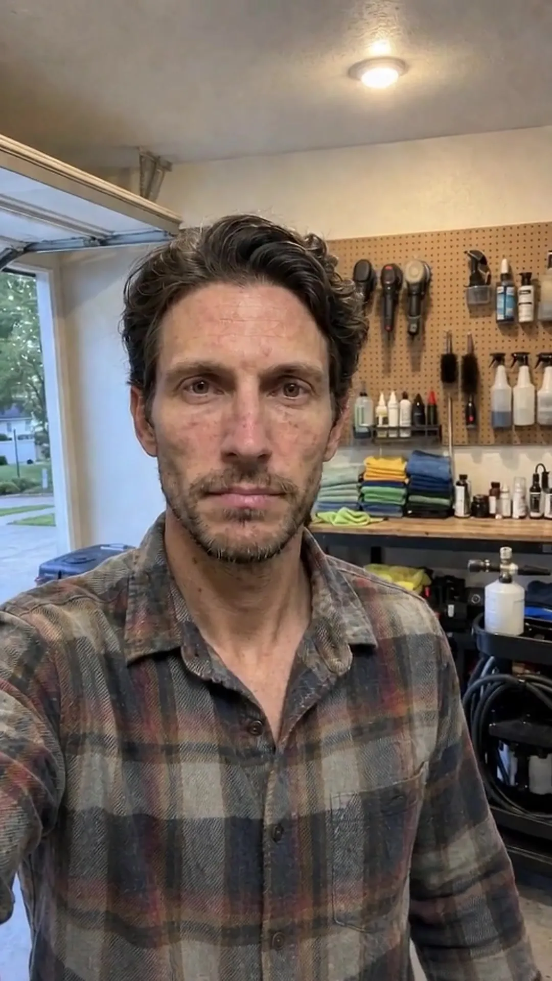 A detailer in a plaid shirt in his garage, a pegboard of brushes and bottles on the wall behind him.