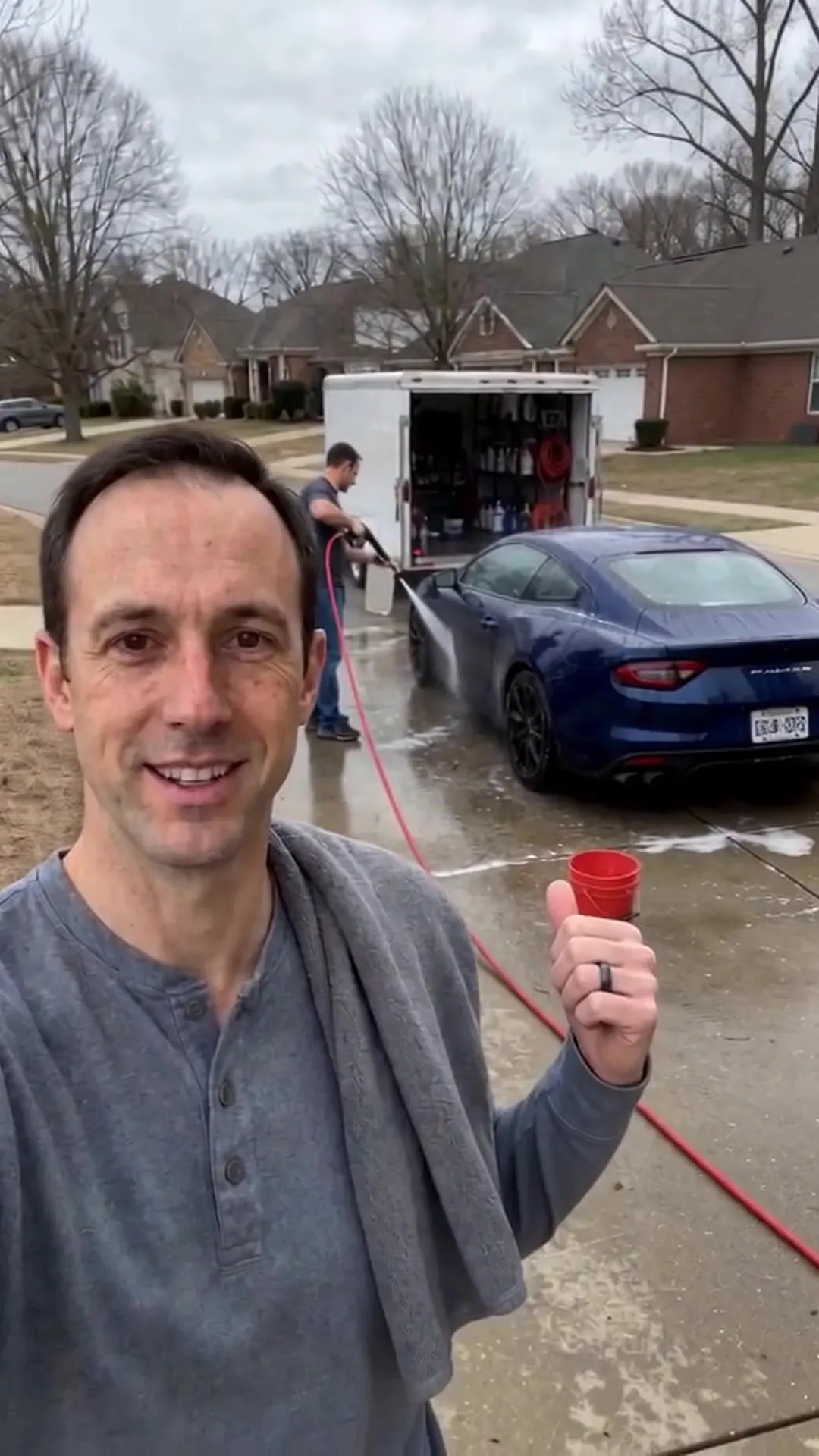 A detailer giving a thumbs up on a wet driveway while a crew member rinses a blue car beside an open trailer.