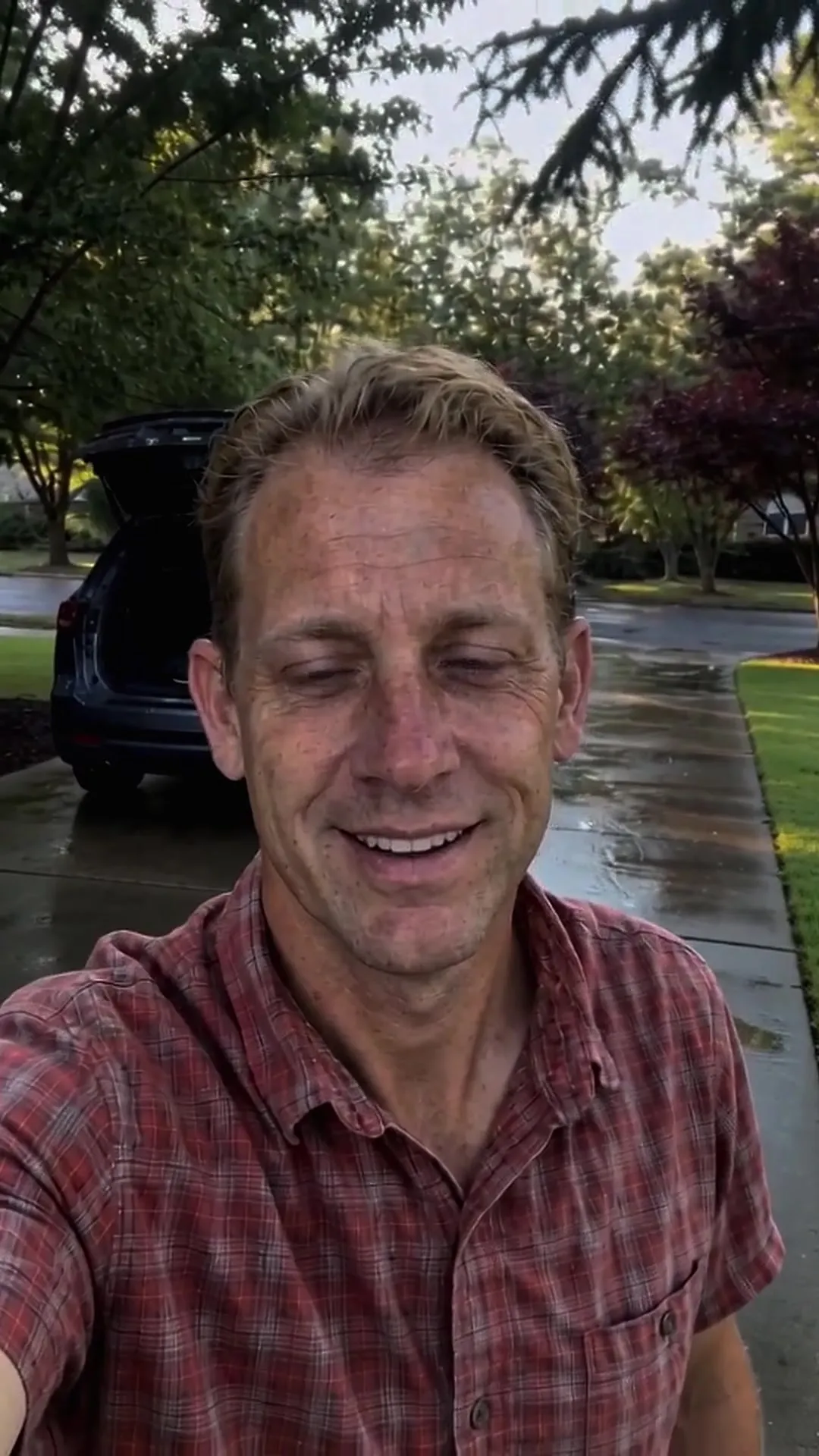 A smiling detailer in a red plaid shirt on a wet driveway, the open tailgate of an SUV behind him.